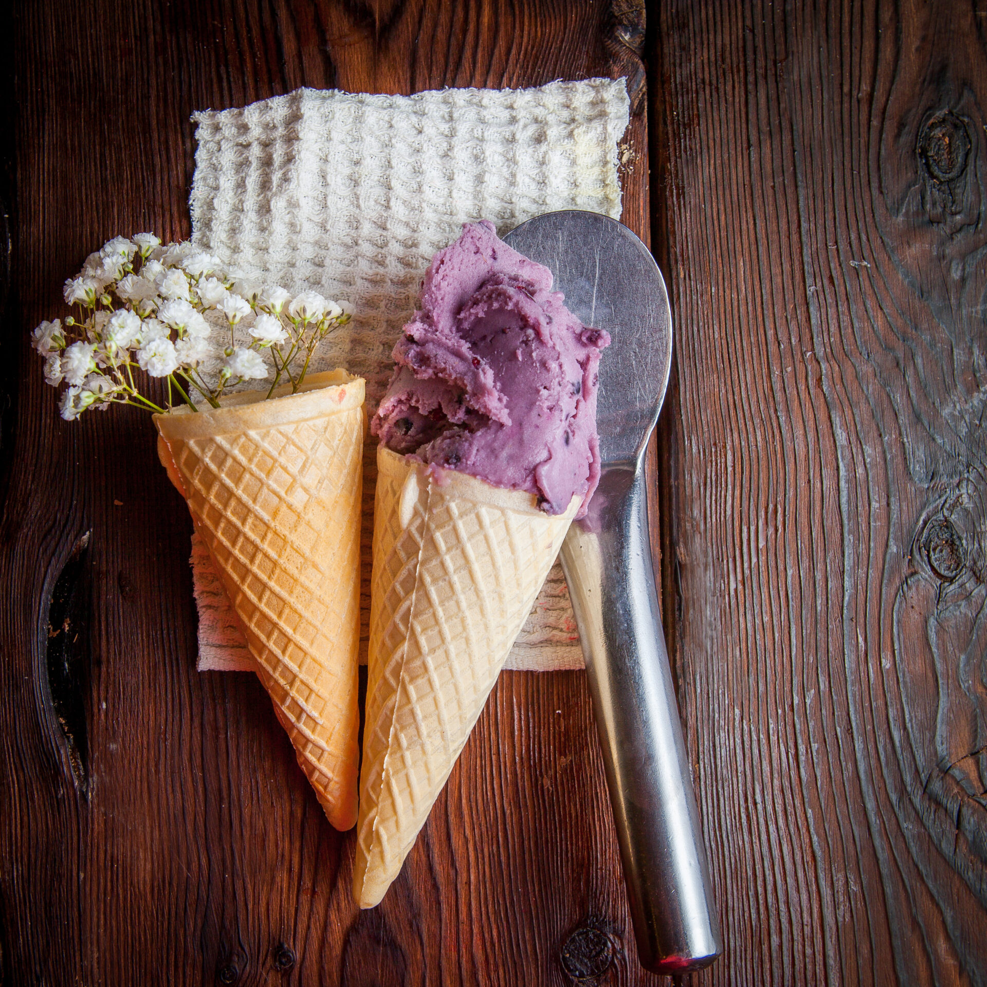 Top view waffle cones with blueberry ice cream and gypsophila and ice cream spoon in rag napkins on wood background | Atelier dell'albergo